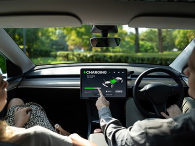 A photo taken inside an EV showing a driver and passenger in the front seat, the driver is pointing to the vehicle's charging screen.